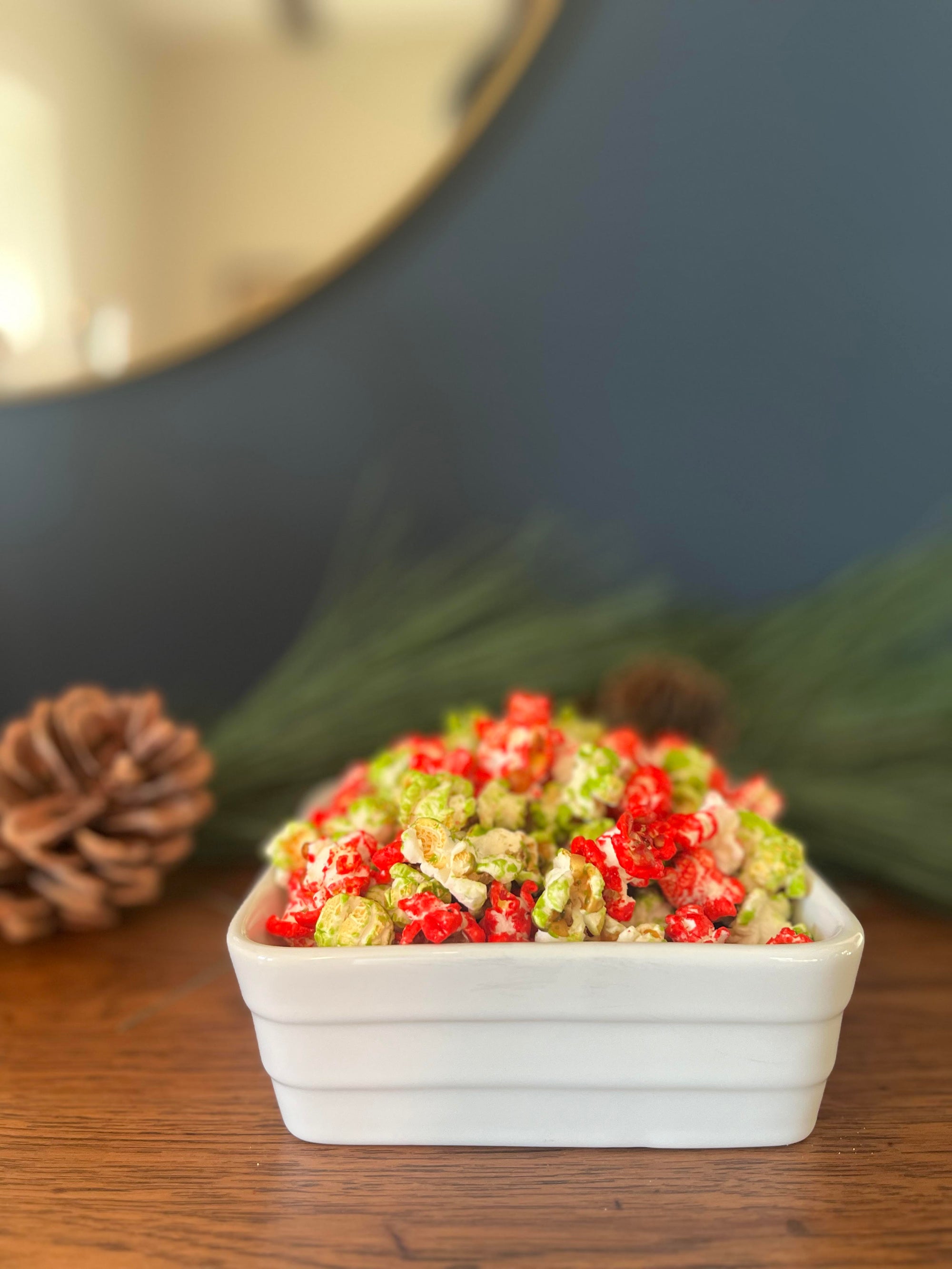 White bowl filled with Bytown Gourmet Popcorn Holiday festive Crunch which consists of red and green candied popcorn sitting on wooden surface with greenery behind
